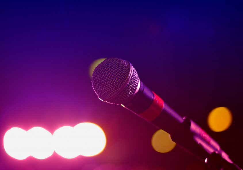 Ein Mikrofon steckt auf einem Mikrofonständer. Im Hintergrund sind Scheinwerfer zu sehen.
